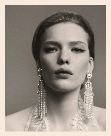Close-up portrait of a stylish woman wearing crystal earrings and elegant makeup, set against a grey background in muted tones.