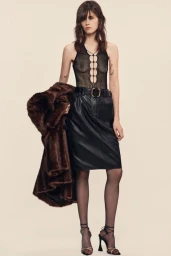 Model in sheer black laced top and leather skirt holding a brown faux fur coat against a seamless light background