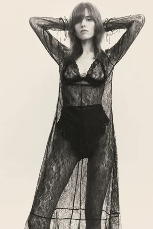 Model posing with hands behind head wearing a sheer black lace long dress over black lingerie against a pale neutral background.