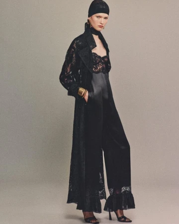 Model in a black lace duster coat, satin jumpsuit, headwrap, and gold cuff bracelet posing against a neutral studio background.