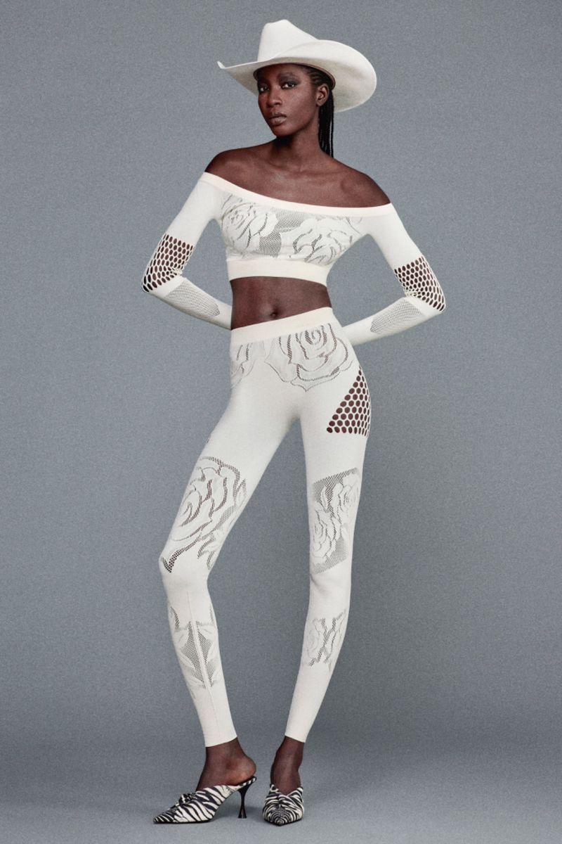 Full-length shot of Black female model posing in off-the-shoulder crop top, leggings, cowboy hat, and heeled mules in a studio setting.