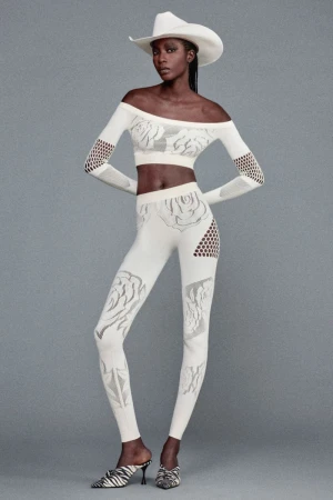 Full-length shot of Black female model posing in off-the-shoulder crop top, leggings, cowboy hat, and heeled mules in a studio setting.