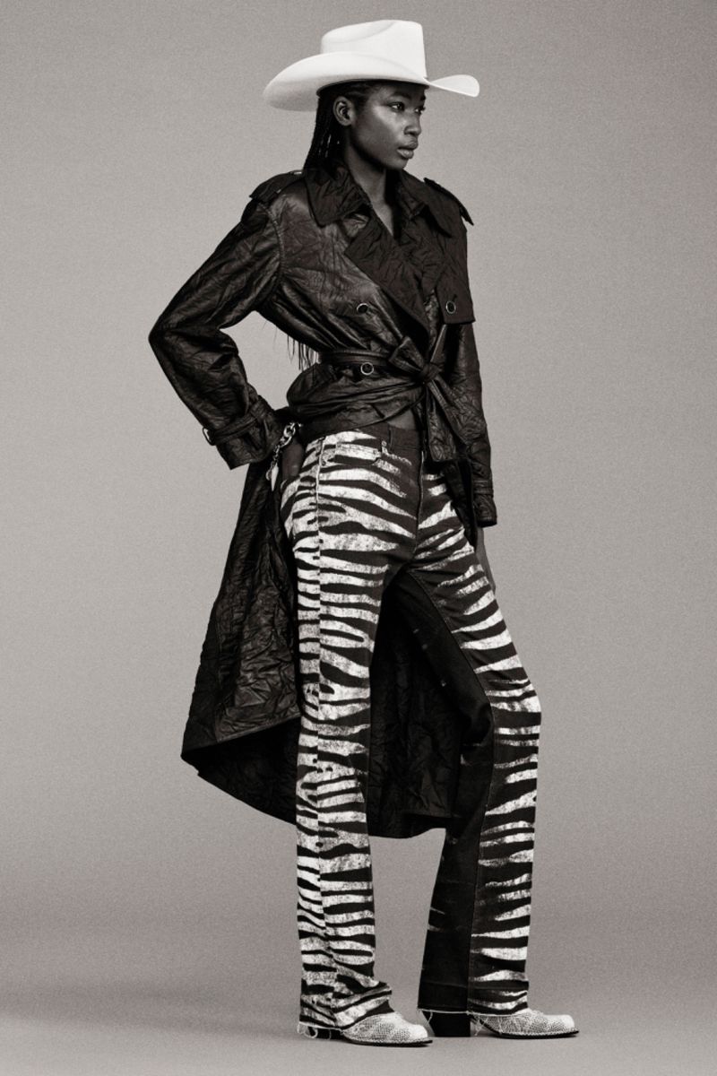 Fashionable woman in zebra-print pants, a dark trench coat, and a cowboy hat stands in a studio setting, showcasing a unique style.