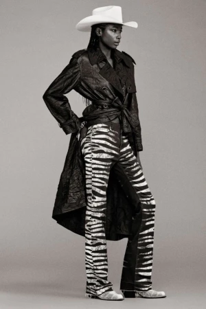 Fashionable woman in zebra-print pants, a dark trench coat, and a cowboy hat stands in a studio setting, showcasing a unique style.