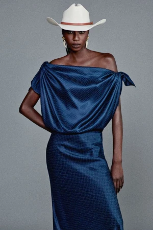 Model wearing an off-the-shoulder draped navy satin dress and a white cowboy hat against a studio grey backdrop.
