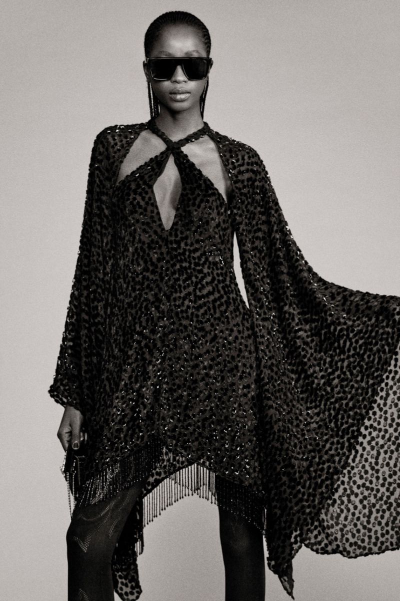 Black and white fashion shot: A female model showcases a dark, semi-sheer dress with fringed hem and cape sleeves, wearing sunglasses.