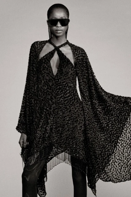 Black and white fashion shot: A female model showcases a dark, semi-sheer dress with fringed hem and cape sleeves, wearing sunglasses.