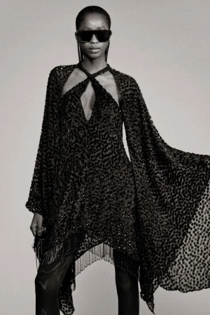 Black and white fashion shot: A female model showcases a dark, semi-sheer dress with fringed hem and cape sleeves, wearing sunglasses.