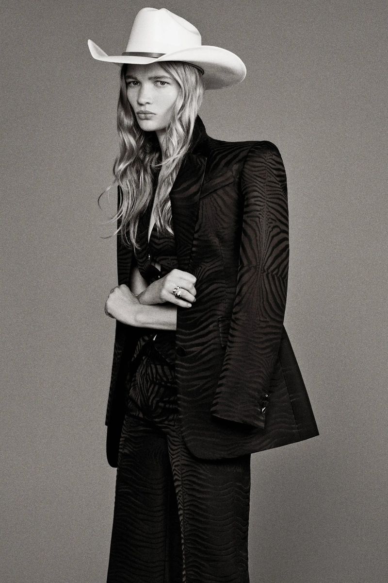 Model wearing a tailored black patterned suit and a white cowboy hat against a grey seamless background.
