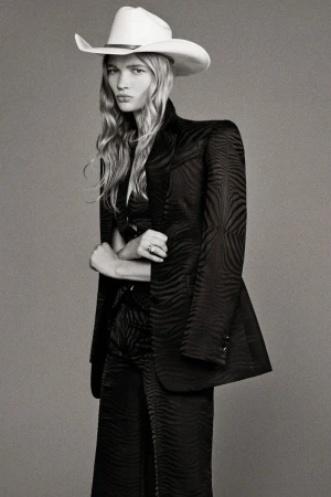 Model wearing a tailored black patterned suit and a white cowboy hat against a grey seamless background.