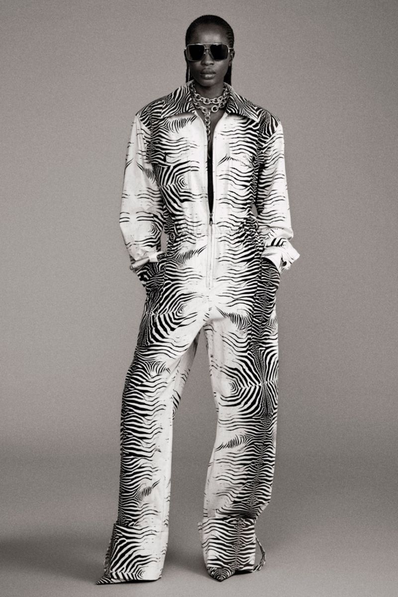 Woman in a black and white zebra-striped jumpsuit, sunglasses, and jewelry, posing in front of a grey backdrop. Full body shot.