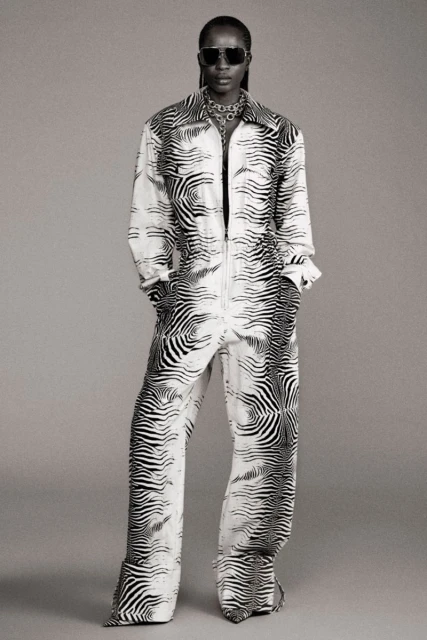 Woman in a black and white zebra-striped jumpsuit, sunglasses, and jewelry, posing in front of a grey backdrop. Full body shot.