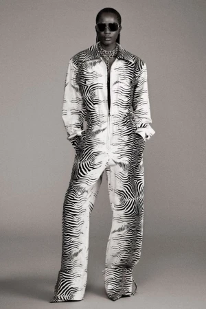 Woman in a black and white zebra-striped jumpsuit, sunglasses, and jewelry, posing in front of a grey backdrop. Full body shot.