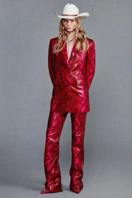 Fashion shot of a female model wearing a red snakeskin-patterned suit and white cowboy hat standing against a grey backdrop.
