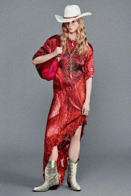 A blonde woman poses in a red floral dress with a white cowboy hat, handbag, necklace, and cowboy boots against a grey background.