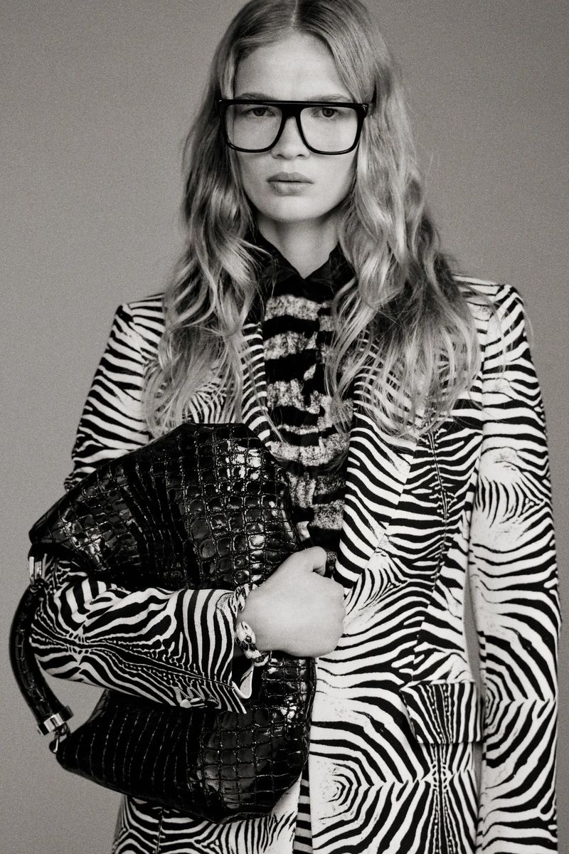A woman poses in a zebra-striped blazer and glasses, holding a black crocodile-skin bag against a gray background in a studio shot.