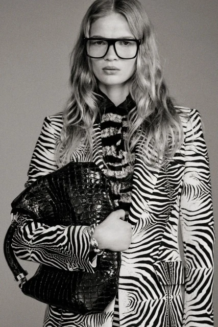 A woman poses in a zebra-striped blazer and glasses, holding a black crocodile-skin bag against a gray background in a studio shot.