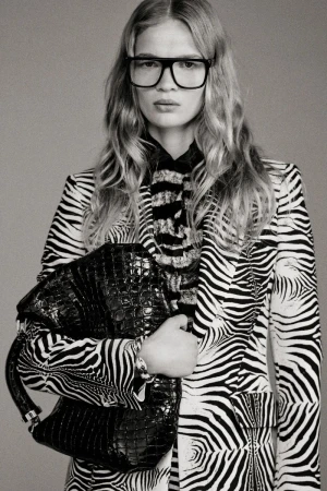 A woman poses in a zebra-striped blazer and glasses, holding a black crocodile-skin bag against a gray background in a studio shot.