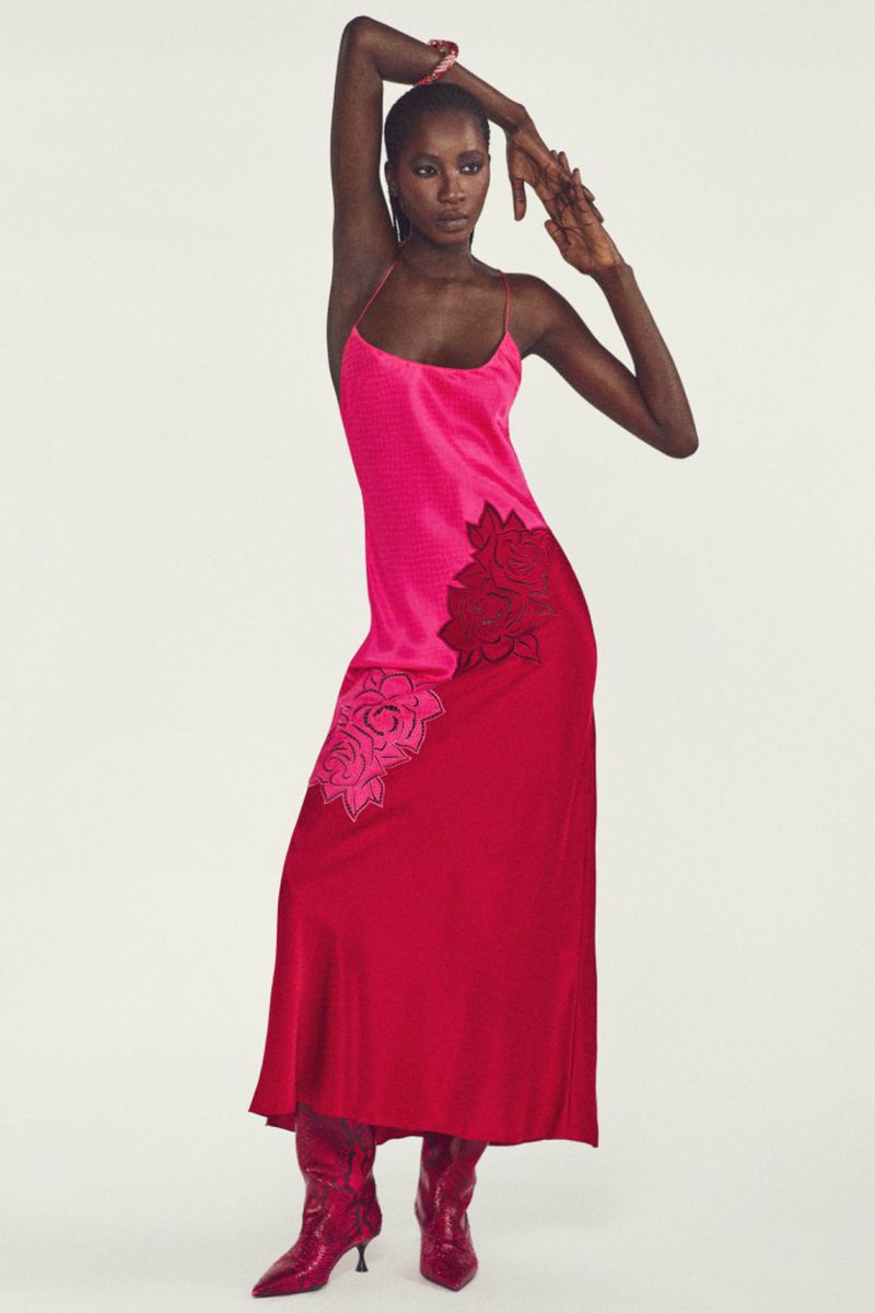 Elegant Black woman poses gracefully in a vibrant pink and red dress with floral accents, paired with matching pink pointed-toe boots on white.