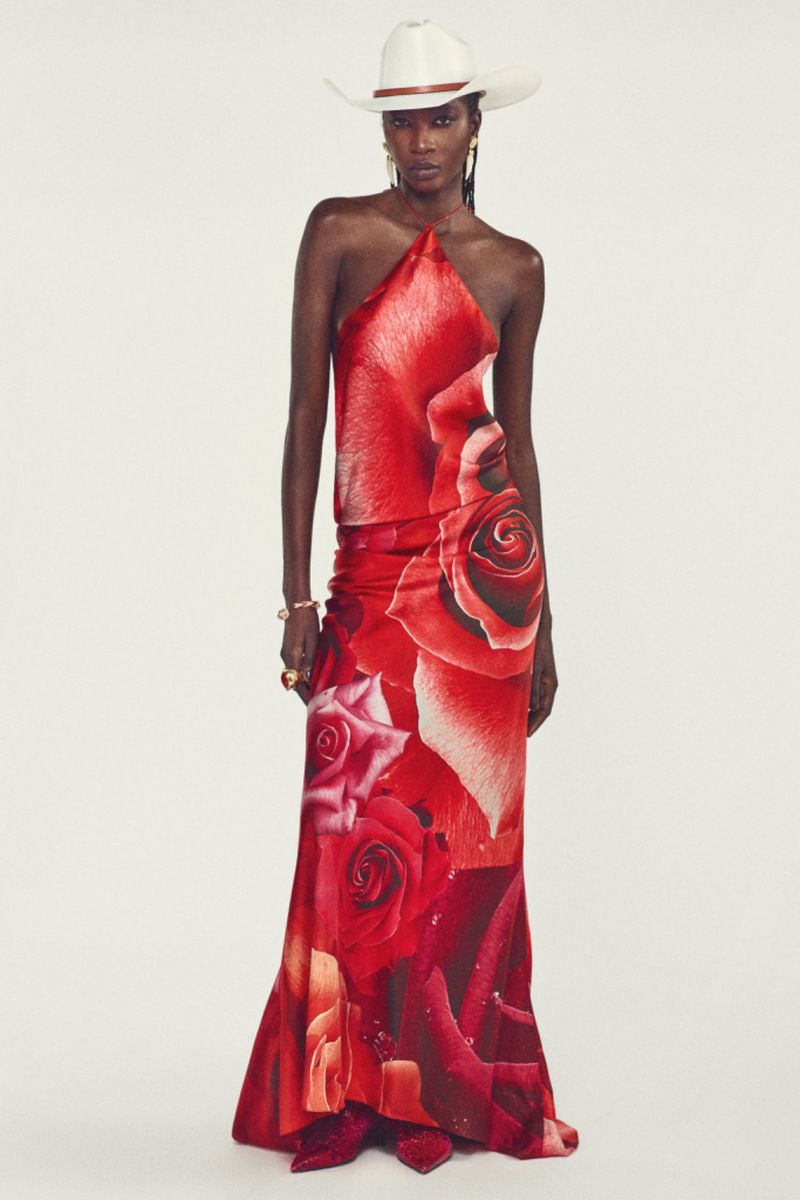 Woman in halter dress with red rose print and white cowboy hat in a fashion studio shoot. Full length shot on white backdrop.