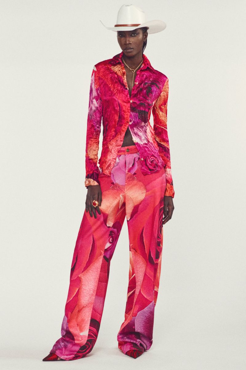 An adult female with dark skin stands against a white backdrop, wearing a floral-patterned outfit and a white cowboy hat.