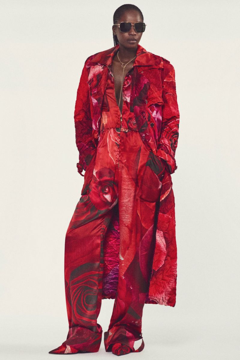 Model wearing dark sunglasses, an all-over red floral print trench coat, matching wide-leg jumpsuit, and pointed shoes on white seamless.