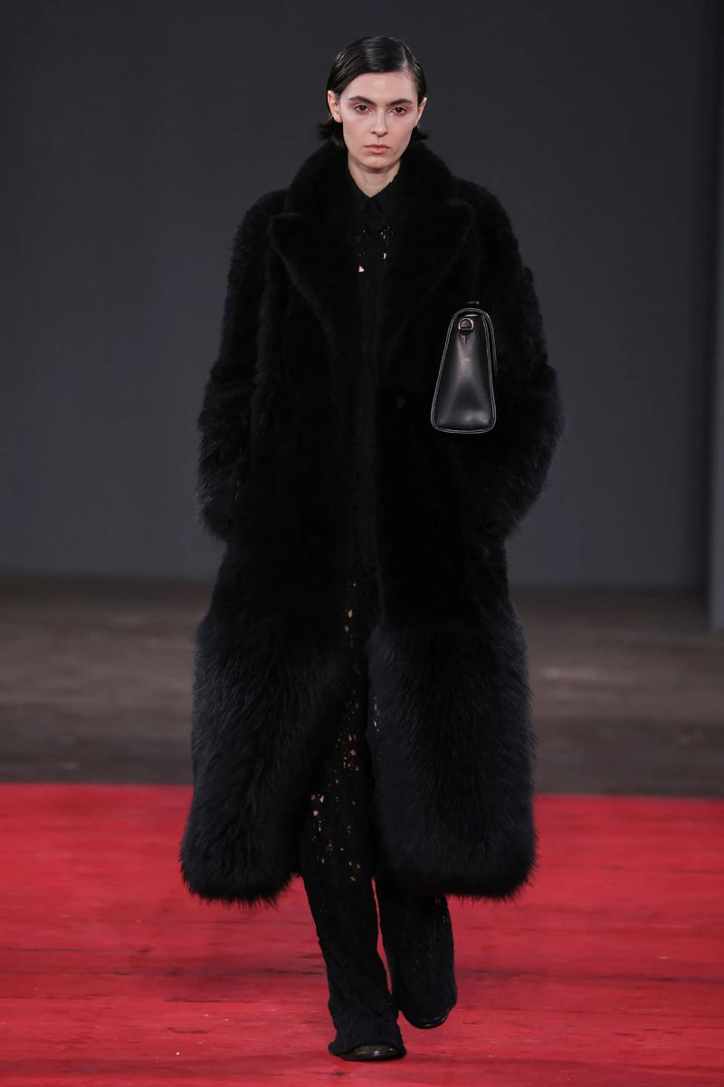 Runway fashion: A woman in a long black furry coat walks the red floor in an indoor space with a black leather handbag.
