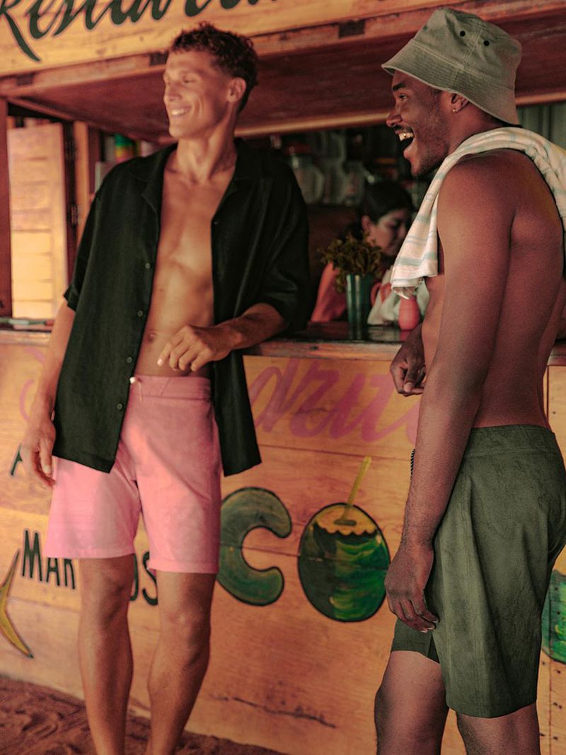 Two men at a rustic beach bar. One man in pink shorts and an unbuttoned shirt smiles while the other in a bucket hat laughs.