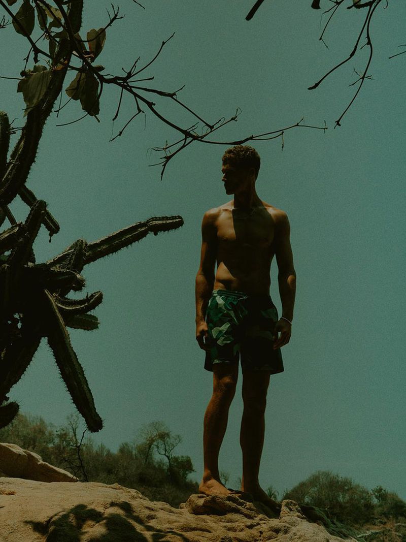 Shirtless model Bram Valbracht standing on rocks beside a cactus in green swim shorts for Zara Man Beachwear.