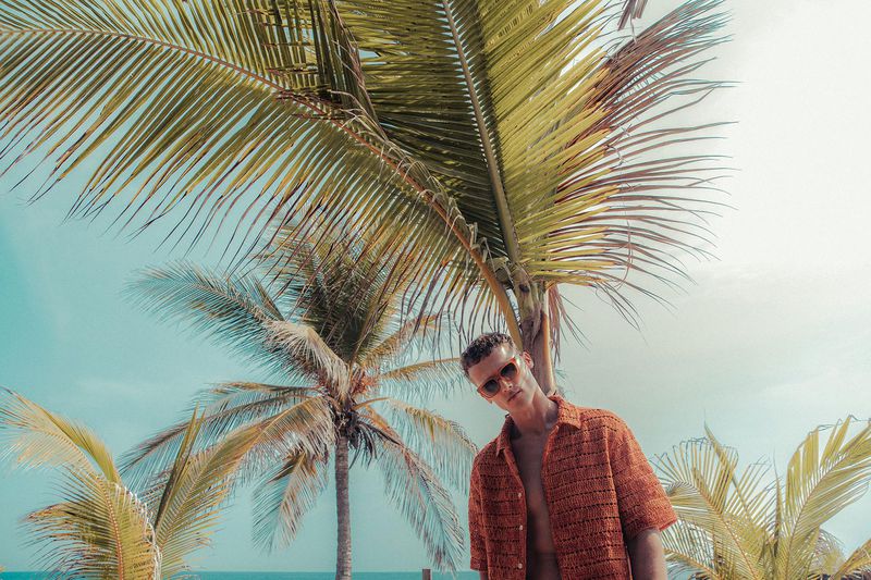 A man poses wearing sunglasses and an orange shirt, under palm trees in a sunny, tropical outdoor setting. Fashion editorial style.