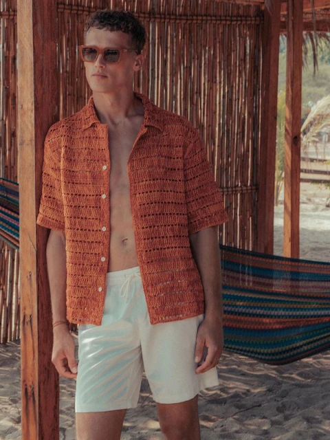 A handsome adult male is featured wearing sunglasses and a crocheted shirt, standing near a hammock at a beachside resort.