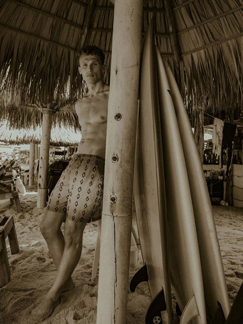 A shirtless man in patterned shorts leans against a bamboo pole, with surfboards nearby under a thatched roof at a beachside hut.