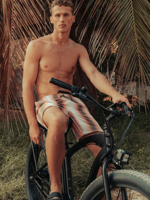 Shirtless man with curly hair is riding an electric bike in tan and peach shorts outdoors in sunlight with palms and greenery.