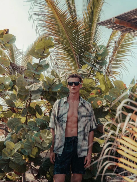 Adult male fashion model poses in front of lush tropical vegetation wearing an open short-sleeve button-up shirt and dark shorts outdoors.