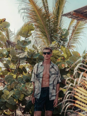 Adult male fashion model poses in front of lush tropical vegetation wearing an open short-sleeve button-up shirt and dark shorts outdoors.