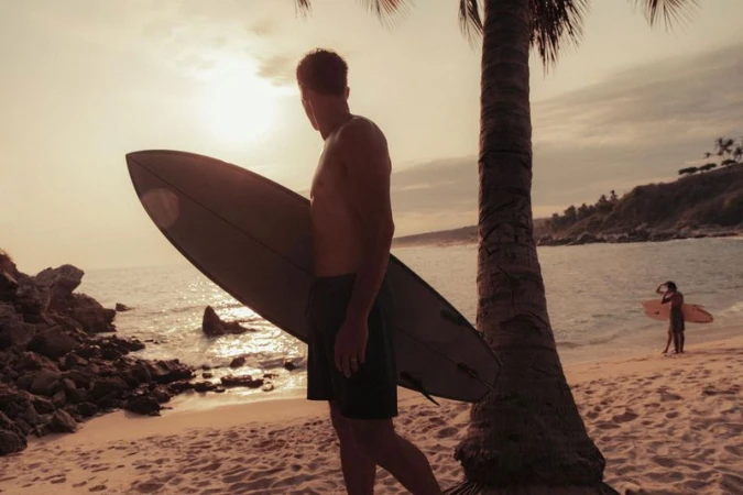 Man stands on a sandy beach near a palm tree, holding a surfboard. Ocean and coast visible in the soft, warm light of dusk.
