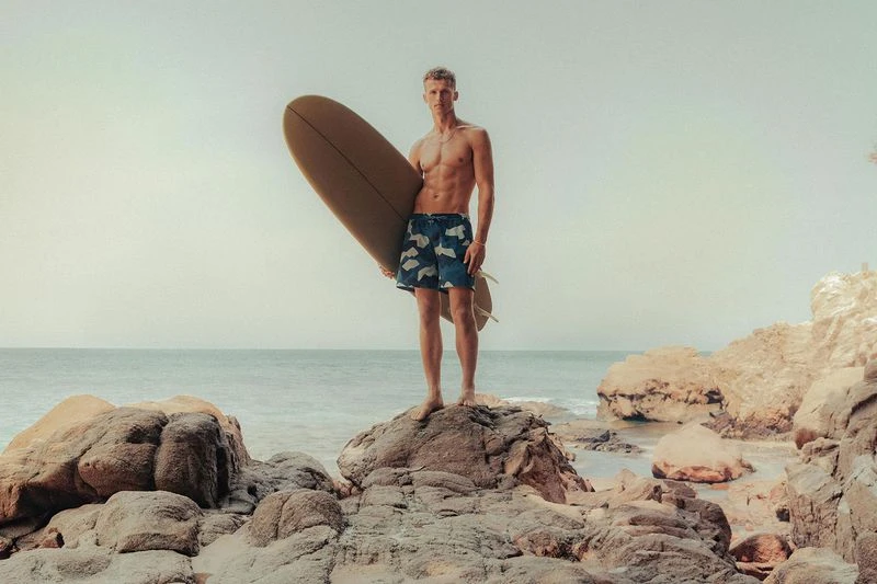 Shirtless man stands on rocks at beach holding surfboard in sunny daylight, ocean background, wearing patterned board shorts.