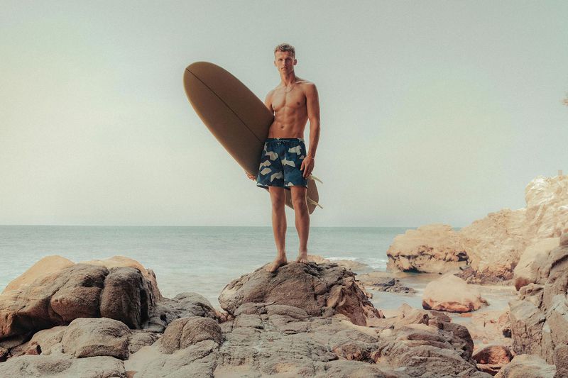 Shirtless man stands on rock formation at beach holding surfboard on sunny day with ocean and sky background.