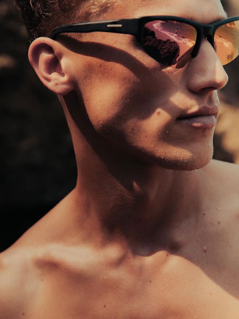 Close-up of a young adult male wearing sunglasses with a landscape reflection, capturing the sunlit skin and stylish eyewear.