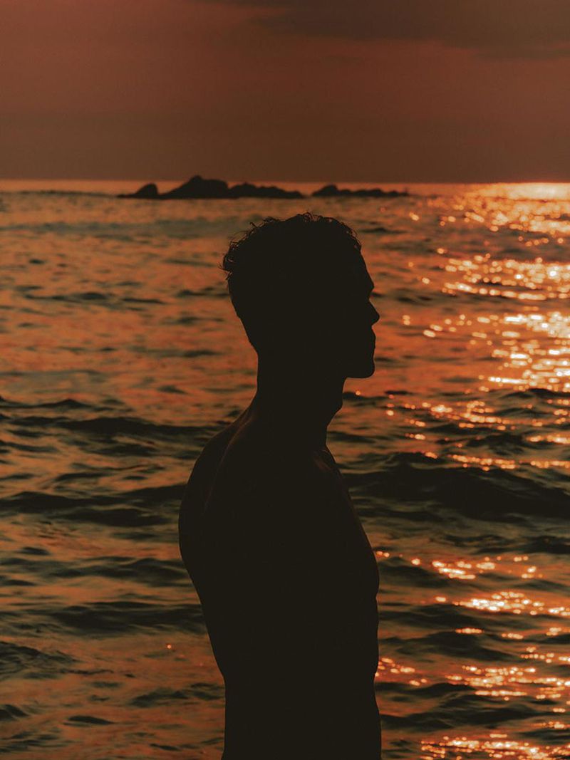 Silhouette of a muscular man standing in the water at sunset with ocean waves, rocks, and sun reflections visible.