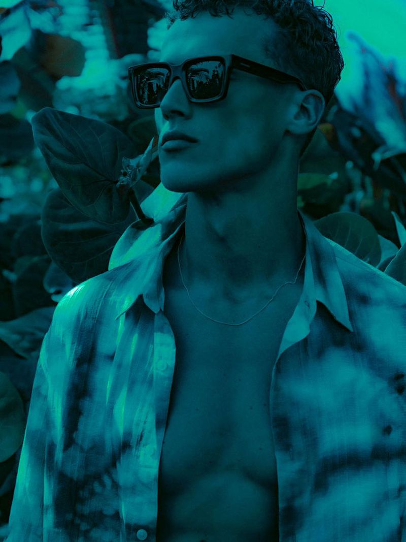 Portrait of a dark-haired adult male with an unbuttoned tie-dye shirt and sunglasses posing outdoors against a leafy background.