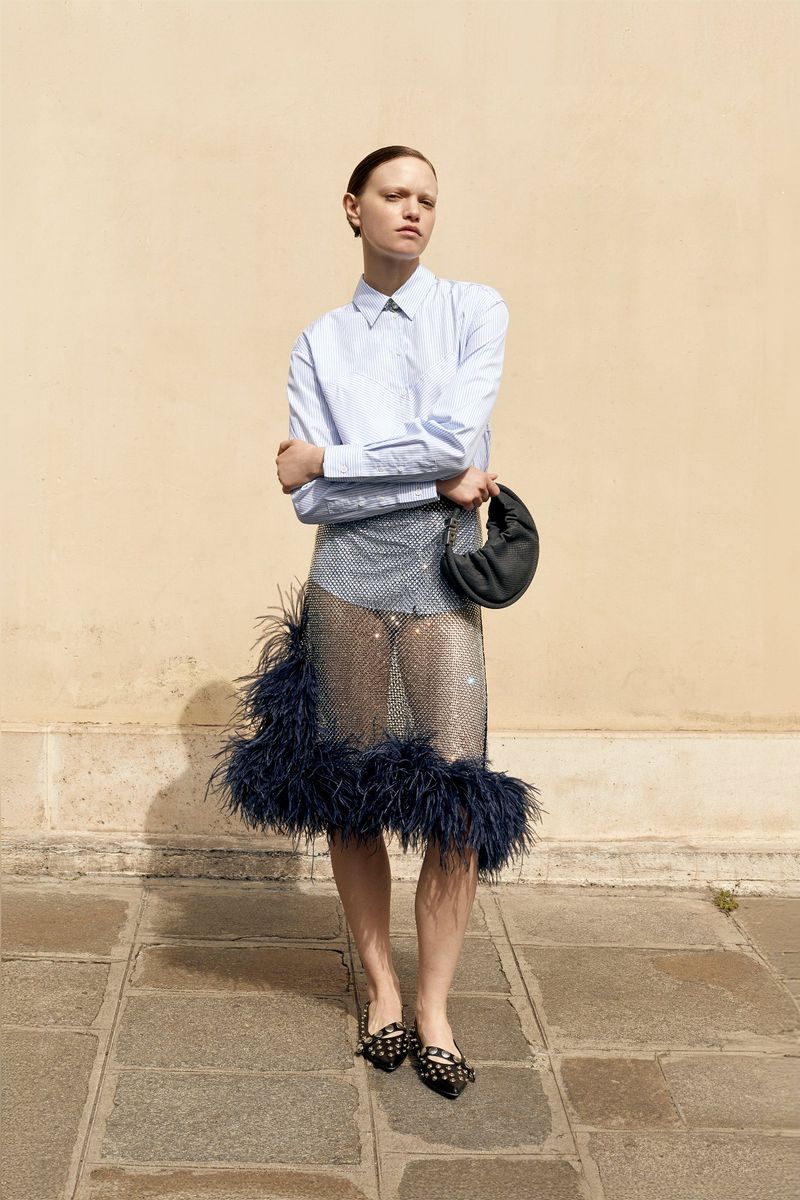 A slender fashion model poses outdoors wearing a light blue button-down shirt, transparent skirt with feather trim, and black heeled shoes.