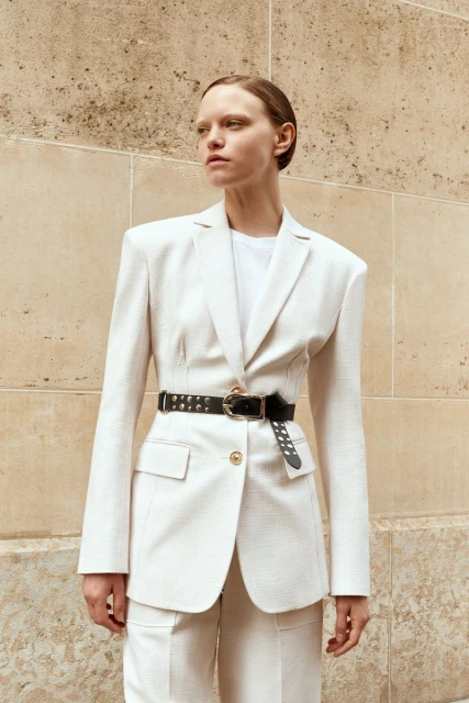Adult woman poses in a light-colored tailored suit and black belt, standing outdoors against a light brown paneled wall on a sunny day.