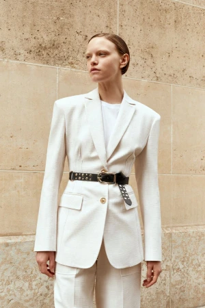 Adult woman poses in a light-colored tailored suit and black belt, standing outdoors against a light brown paneled wall on a sunny day.