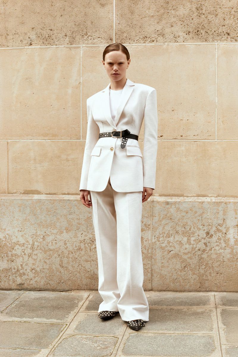 A stylish woman in a white pantsuit with a black belt stands against a textured wall and cobblestone. Fashion shoot, neutral tones.