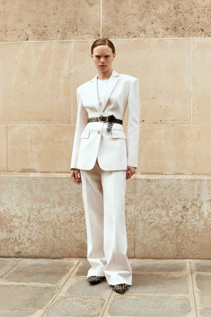 A stylish woman in a white pantsuit with a black belt stands against a textured wall and cobblestone. Fashion shoot, neutral tones.