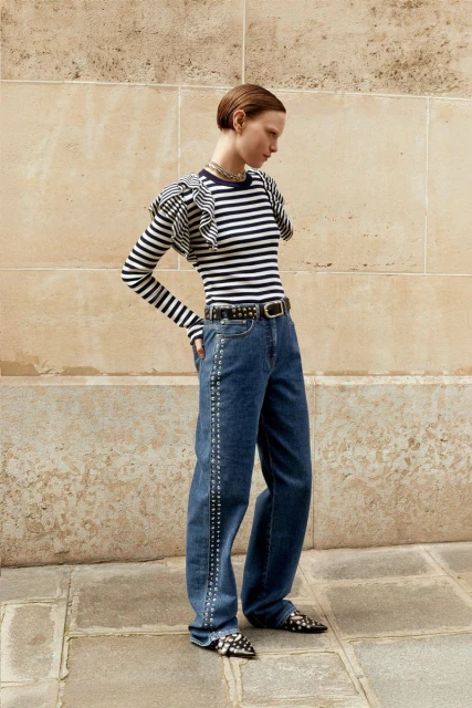 A stylish woman poses outdoors in a striped top, wide-leg jeans with studded details, a studded belt, and pointed-toe shoes for a fashion shoot.