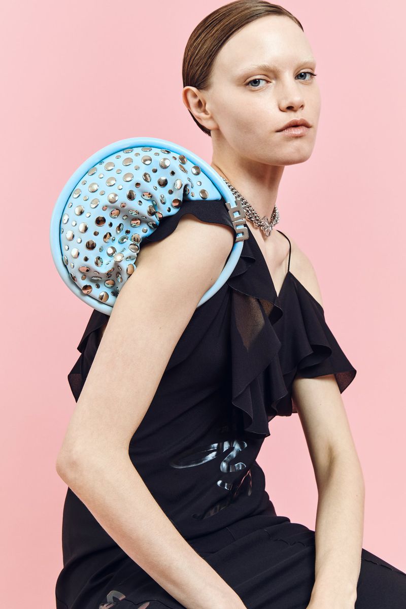 Fashion shot of a woman in a black dress with a unique blue round bag, set against a light pink backdrop, showcasing modern style.