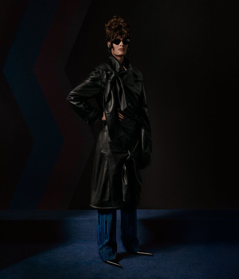 A fashionably styled adult poses in a black leather trench coat and blue wide-legged pants with metal heel accents in a dark studio.