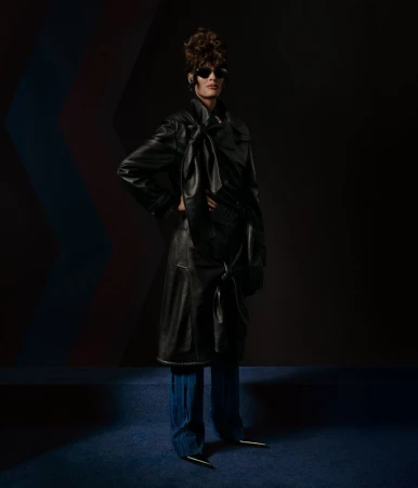 A fashionably styled adult poses in a black leather trench coat and blue wide-legged pants with metal heel accents in a dark studio.